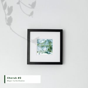 closeup photo of a cherub with moss on its face taken at Mt. View Cemetery in Oakland CA - framed in a square box frame on a gray wall with the shadow of a branch on the wall and the text Cherub #2 / Magic Turtle Studios