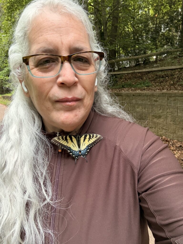 Yo, una mujer de aspecto muy agotado con cabello blanco y gafas, vistiendo una chaqueta marrón. Una hermosa mariposa descansa cerca de mi hombro, habiendo trepado por todo mi cuerpo.